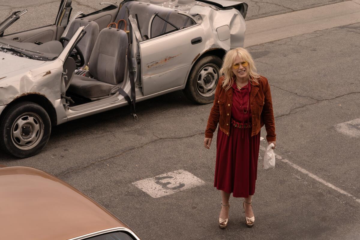 A woman in a maroon dress and jacket stands in front of a beat up silver sedan with no roof.