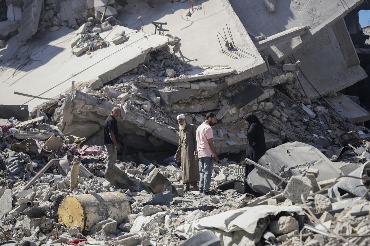 Palestinians stand, dwarfed by massive piles of the rubble of destroyed buildings. 