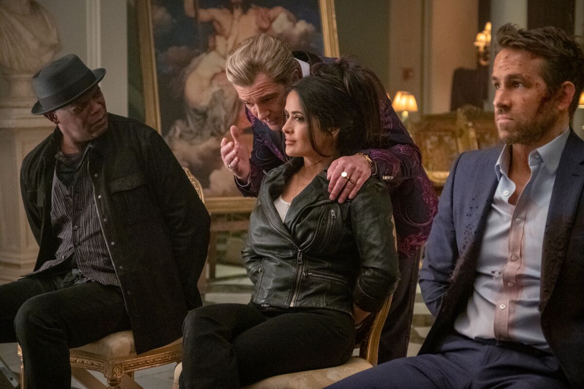 Samuel L. Jackson, Salma Hayek and Ryan Reynolds sit with their hands behind their backs as Antonio Banderas leans in. 