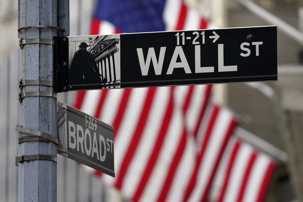 A Wall Street sign is outside the New York Stock Exchange. In the background are American flags.