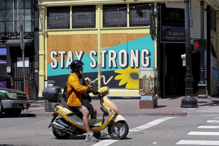 A man riding his scooter waits at the traffic signal on the corner of Fifth Avenue and F Street, where a large mural is painted on wood panels. The panels are fastened to the front entrance to The Tipsy Crow in the Gaslamp Quarter in downtown San Diego.