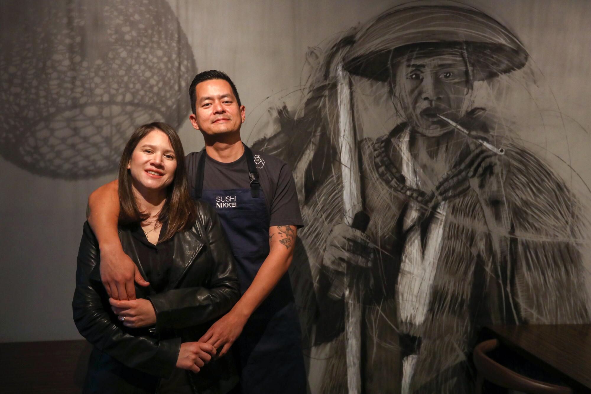 A man stands with his arm around a woman's shoulders, in front of a black-and-white mural of a Japanese farmer