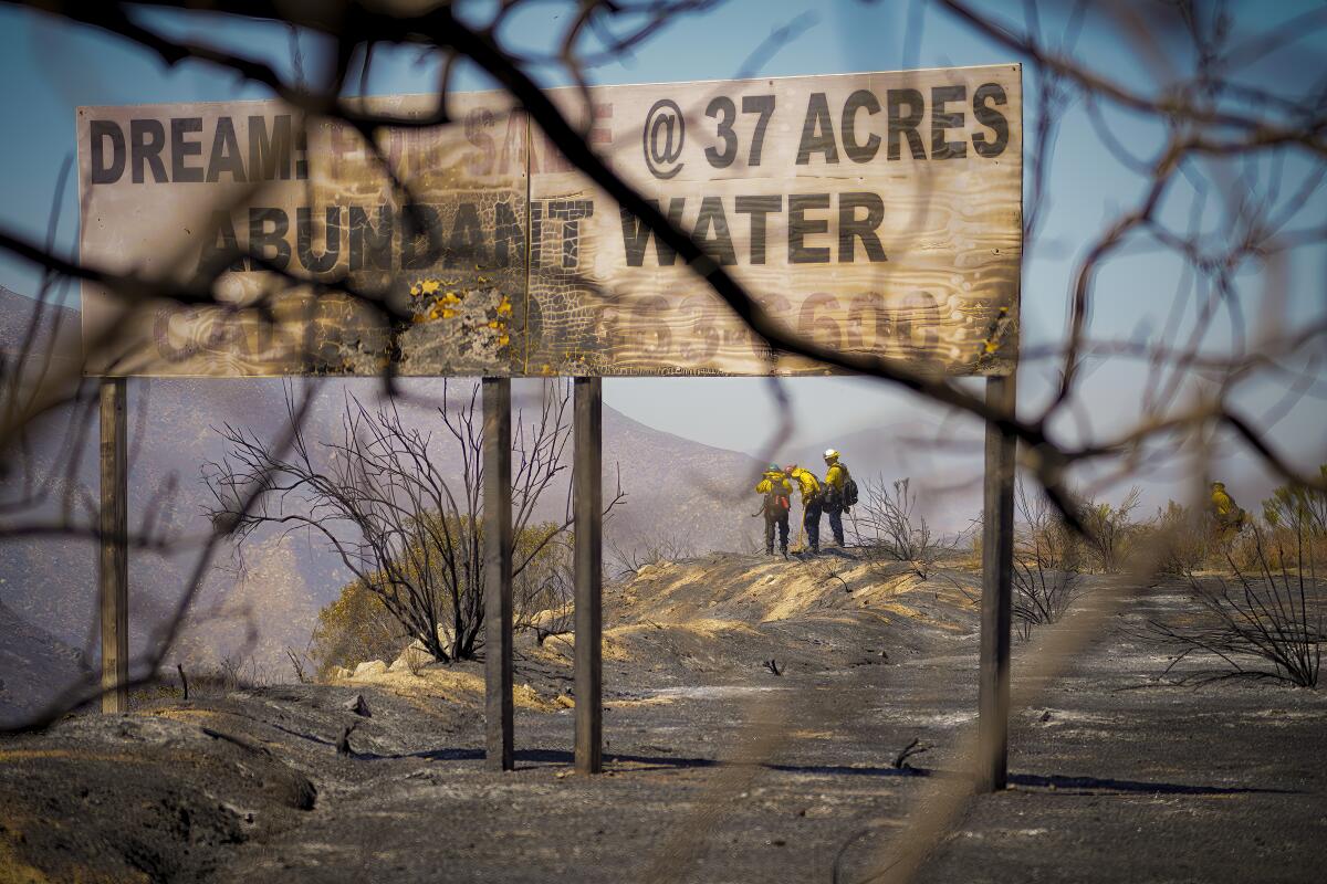 Firefighters stand in a burned area with a charred sign in the foreground