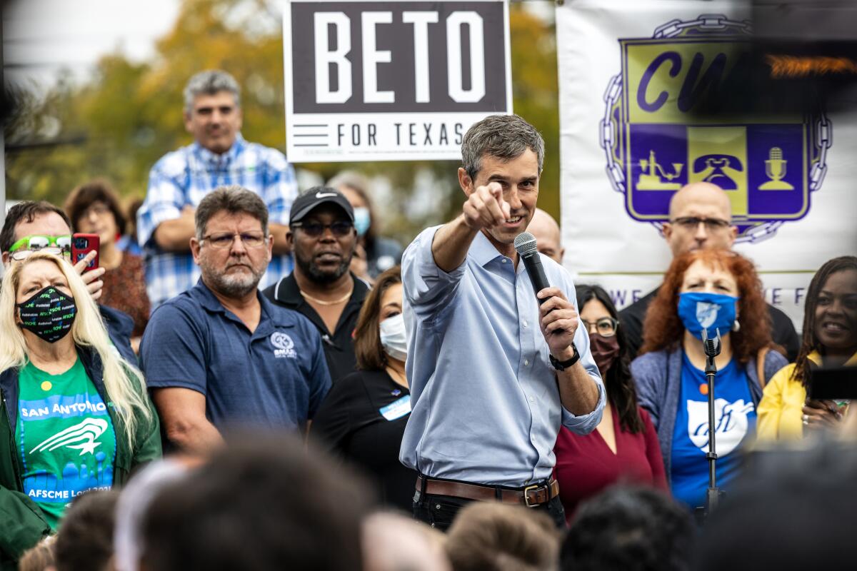 A man in a blue shirt, holding a microphone, points with his other hand as he is surrounded by other people 