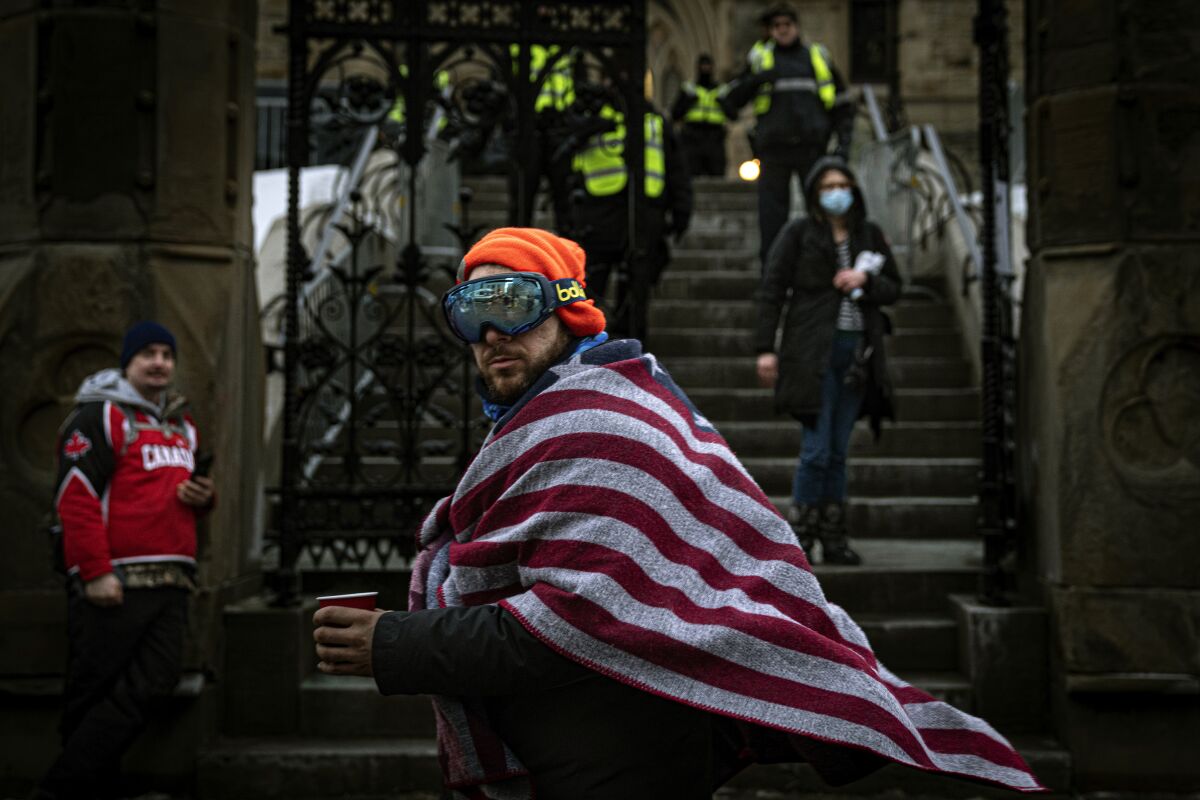 A man wearing an American flag walks by police guarding the Canadian parliament building from demonstrators protesting the country's COVID-19 restrictions, Wednesday, Feb. 16, 2022, in Ottawa, Ontario. The protests in Canada that have blocked border crossings with the U.S. and gnarled trade have been promoted, cheered and funded by American anti-vaccine groups, right-wing activists and conservative elected officials. (AP Photo/Robert Bumsted)