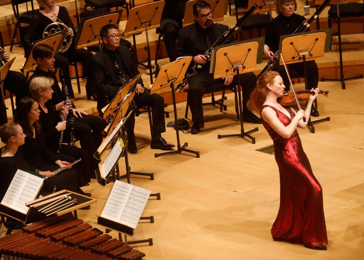 A woman in a red gown plays violin in front of an orchestra