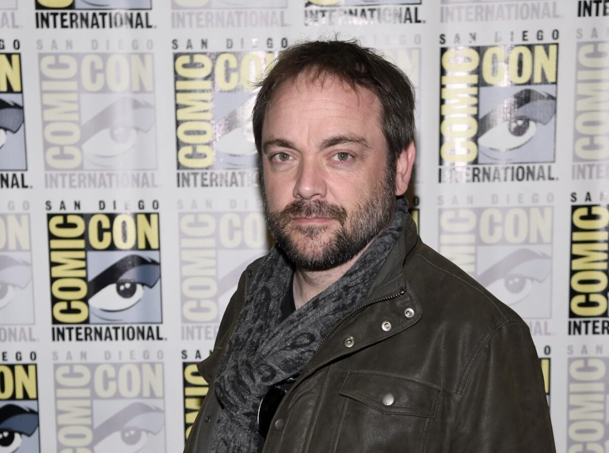 A bearded Mark Sheppard stands in front of a Comic Con backdrop while wearing a jacket and scarf