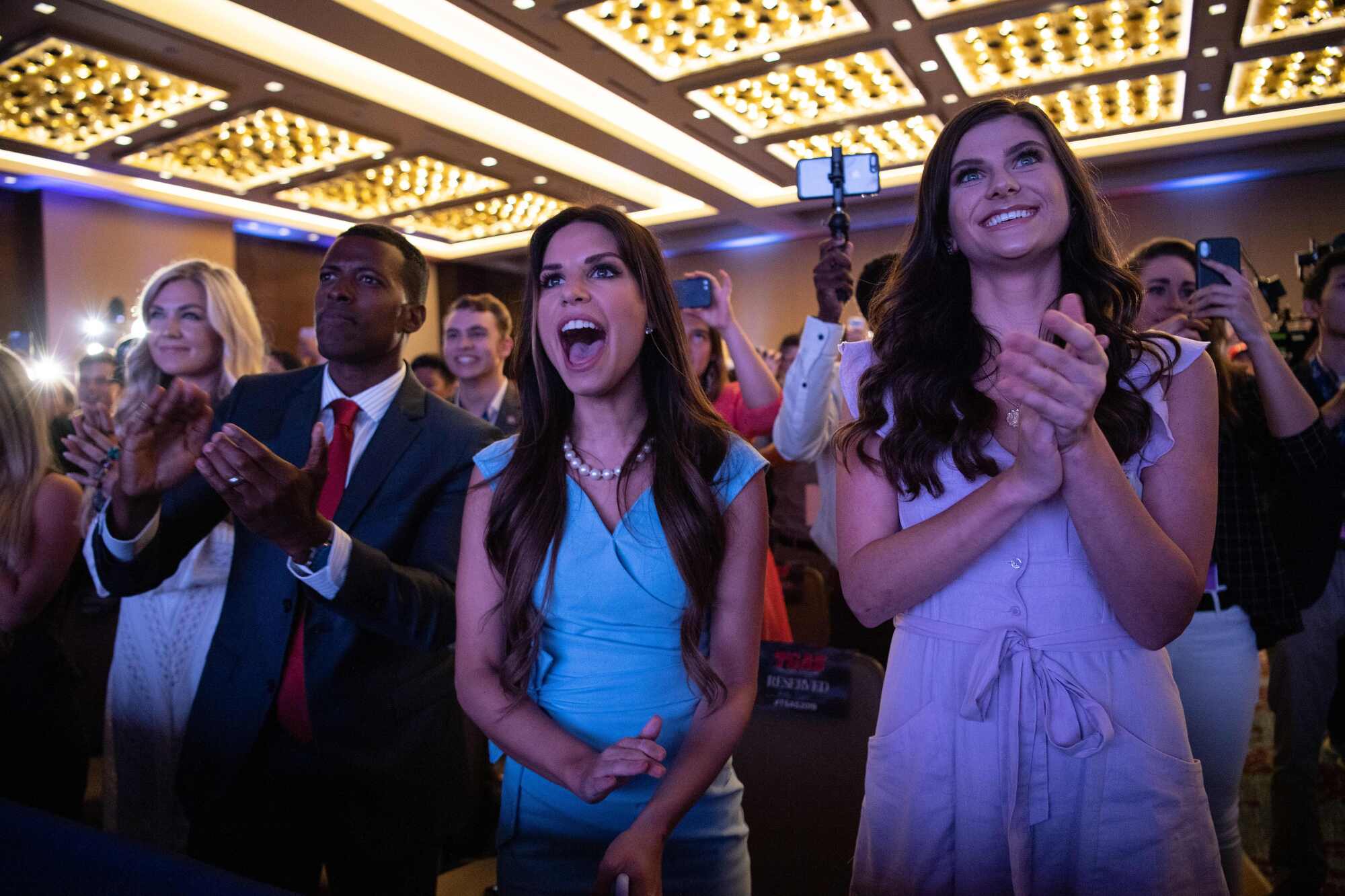 A man in a suit and three women in dresses clap as they stand in front of a crowd in a room