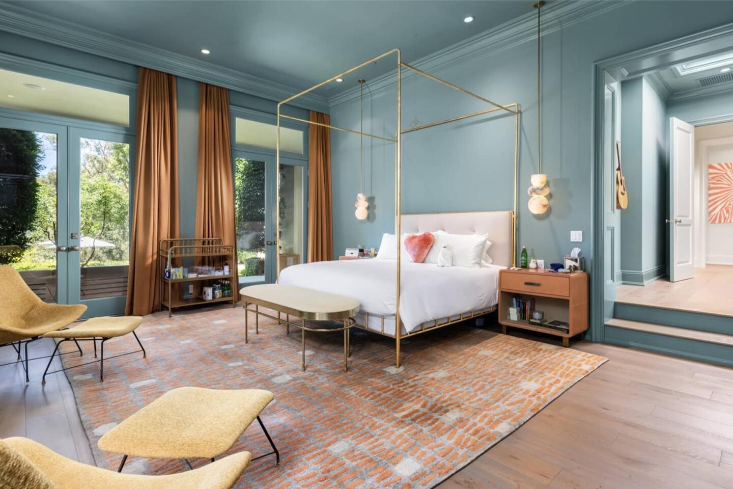 The bedroom is furnished and has bluish-green walls and windows overlooking greenery.