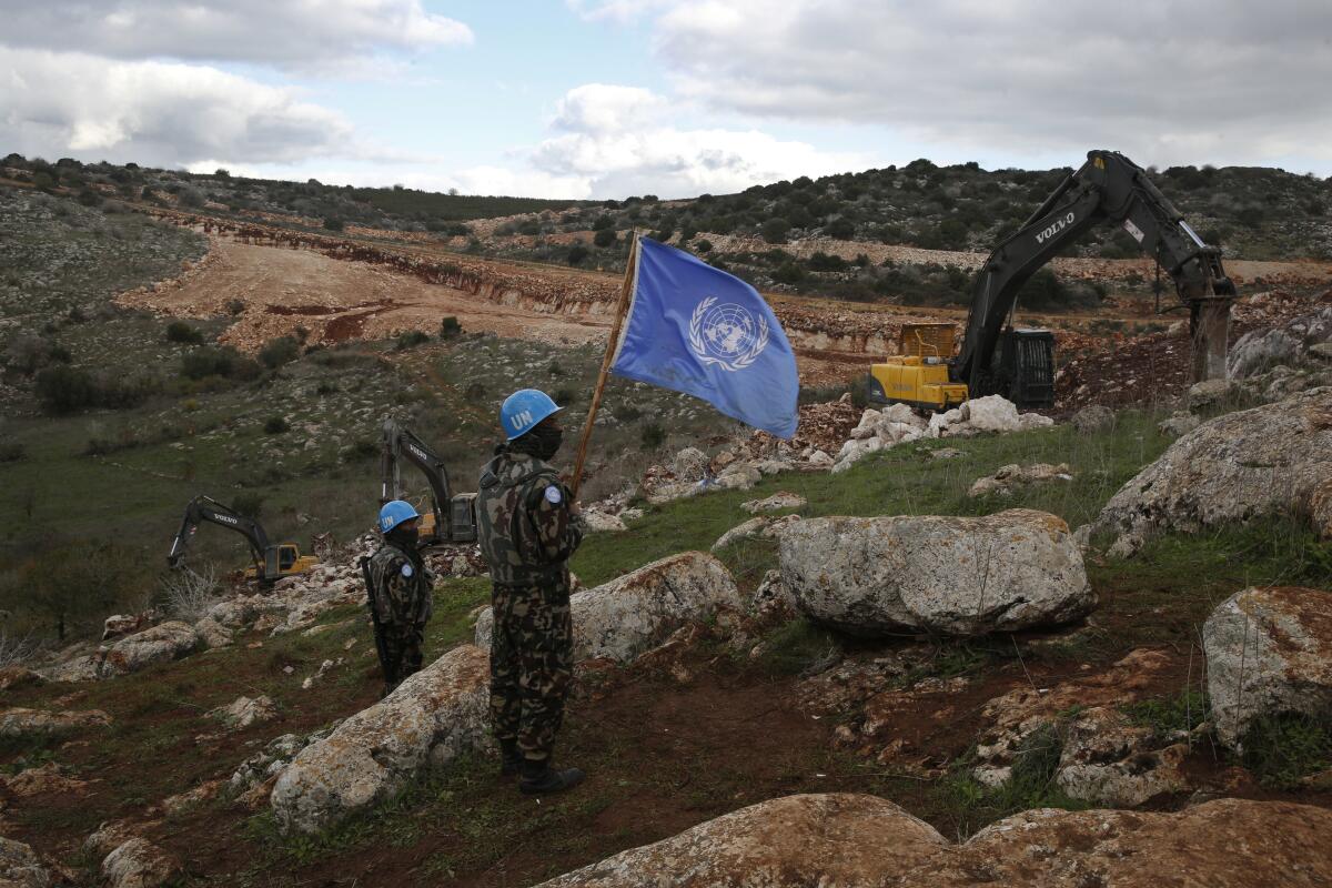 United Nations peacekeepers hold their flag, as they observe an excavator machine working on a rocky hillside.