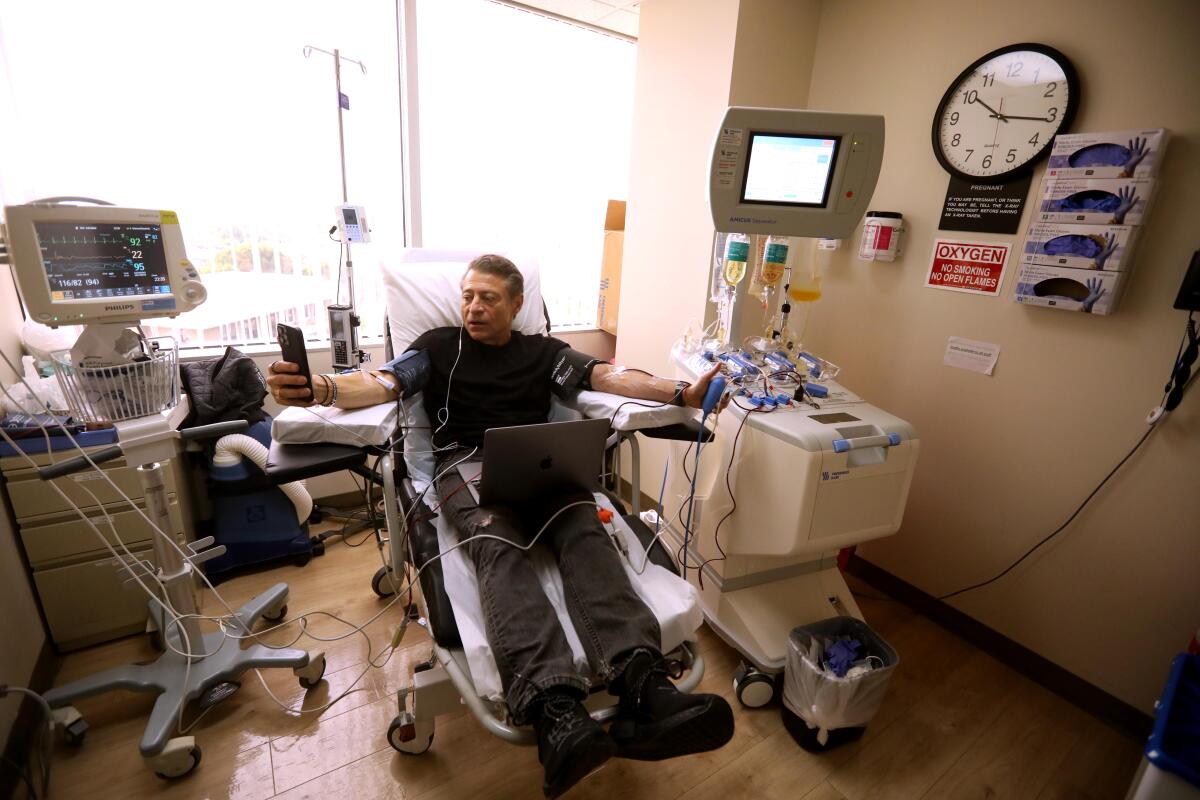 A man sits on a gurney in a medical treatment room receiving intravenous fluids while checking his cellphone