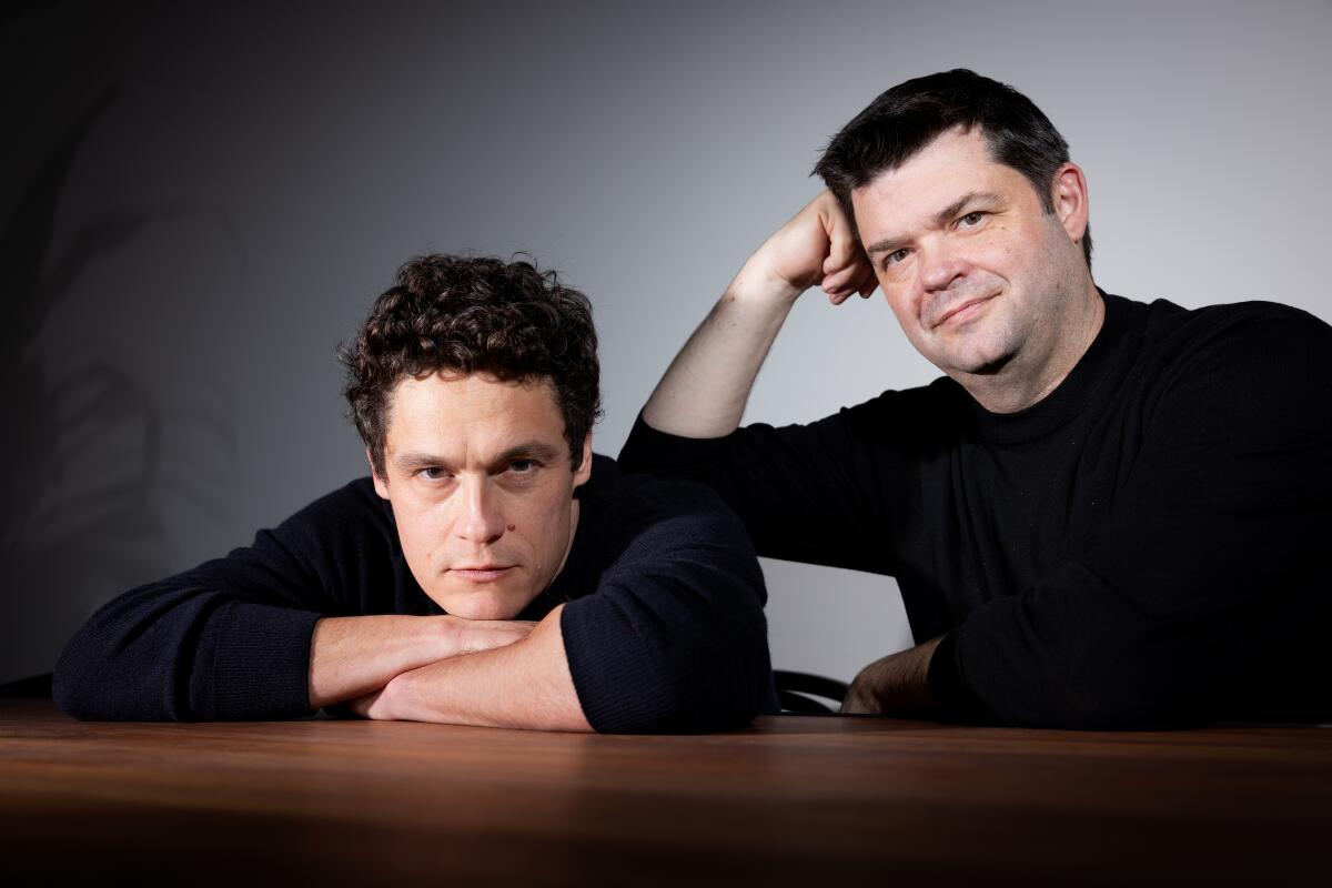 Two men in long-sleeved black T-shirts pose for a portrait, one leaning on a table, the other leaning on the first.