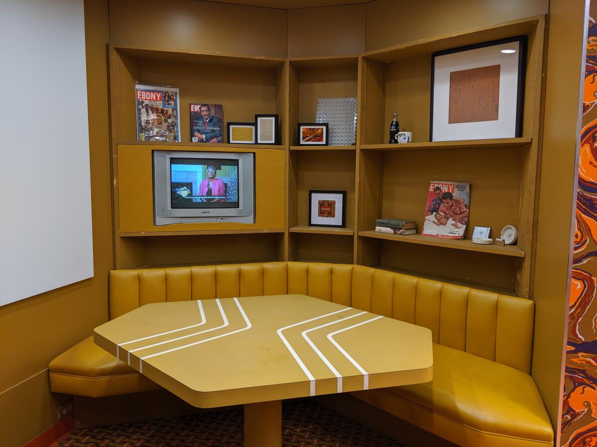 A geometric table in gold with diner-style banquette seating in gold. On shelves are framed artwork and a small TV.