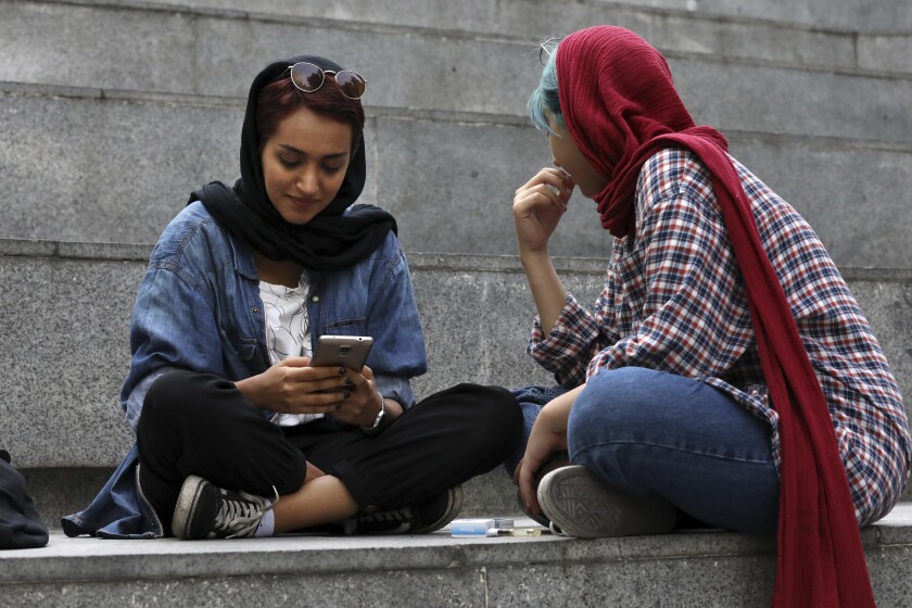 Two Iranian women, one on her cellphone, sit on steps outside a Tehran shopping mall in 2019.