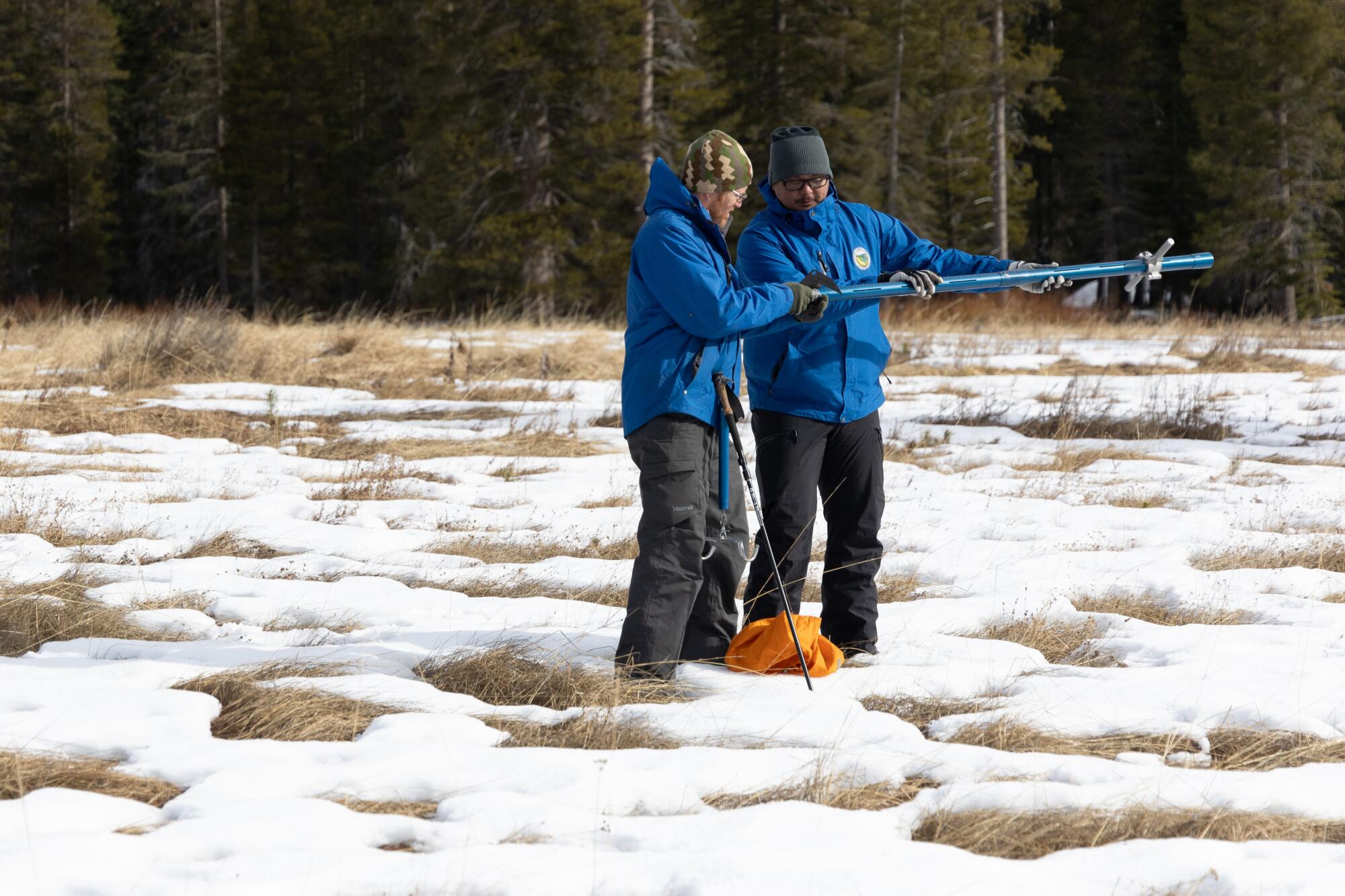 Two men in blue parkas stand in a field of snow and weeds holding a tube used to measure snow depth.