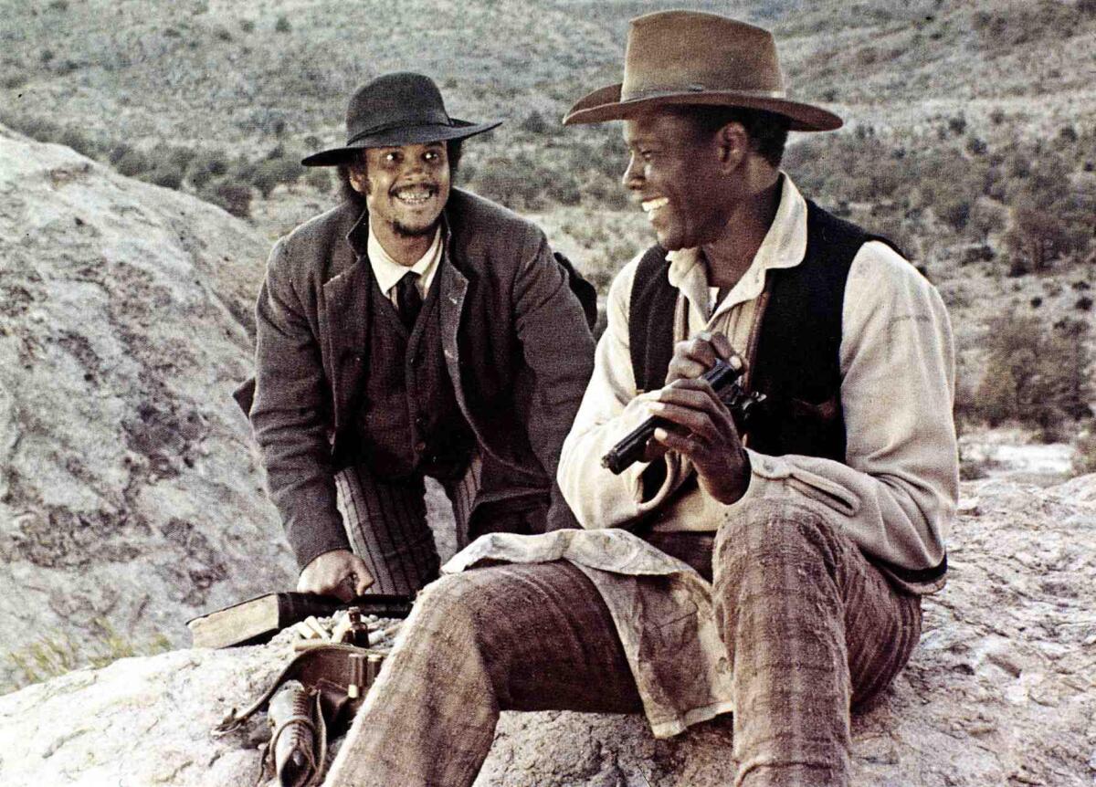 Harry Belafonte grins as Sidney Poitier grins while sitting on a rock.