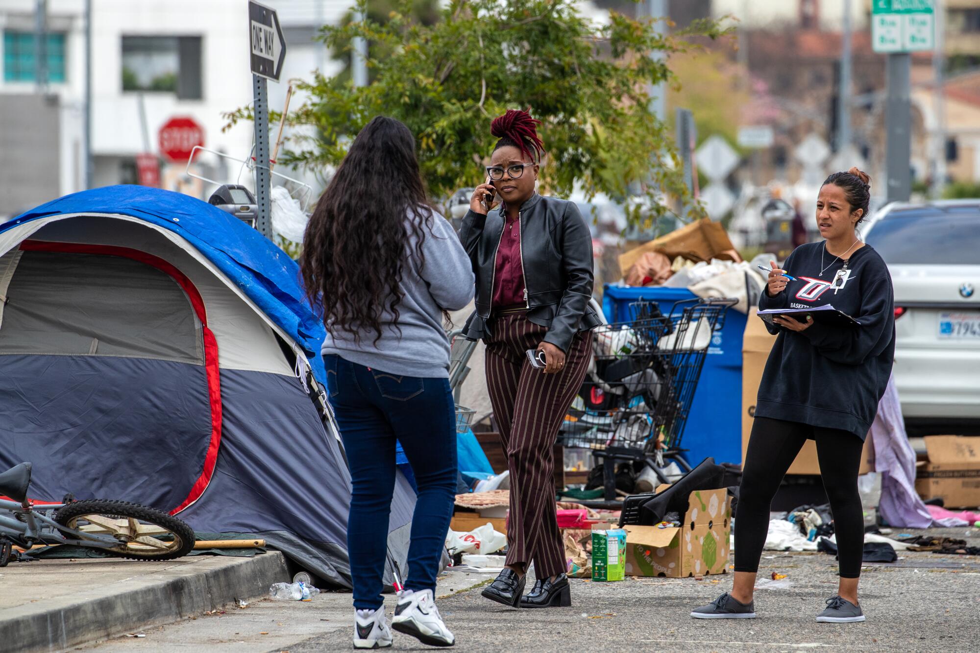 Two women speak to a third at an encampment on San Vicente Boulevard where tents and belongings cover the sidewalk