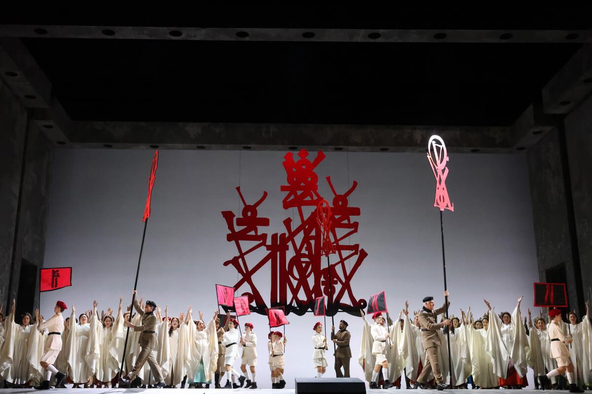 Many actors dressed in white onstage with a large red sculpture above them 