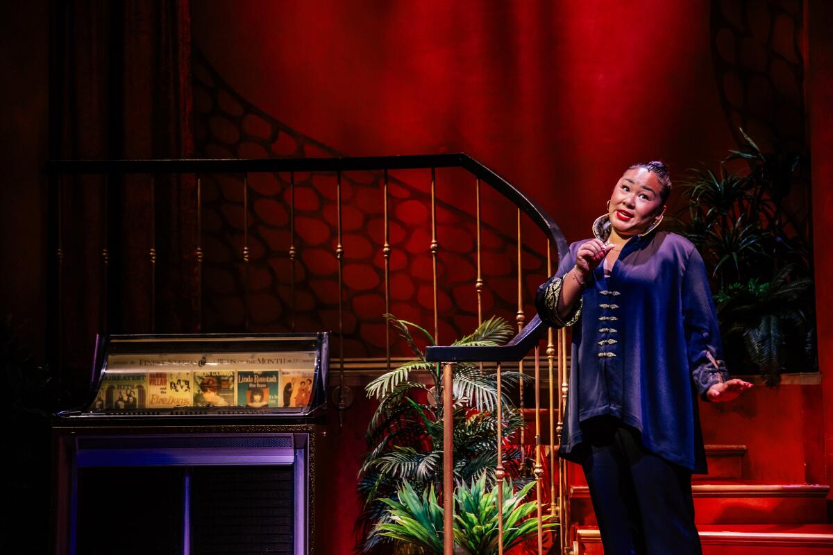 Sara Porkalob stands on a staircase next to an upright piano, her head tilted