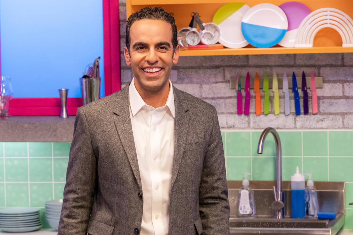 A smiling man in a tweed jacket stands in a colorful kitchen.
