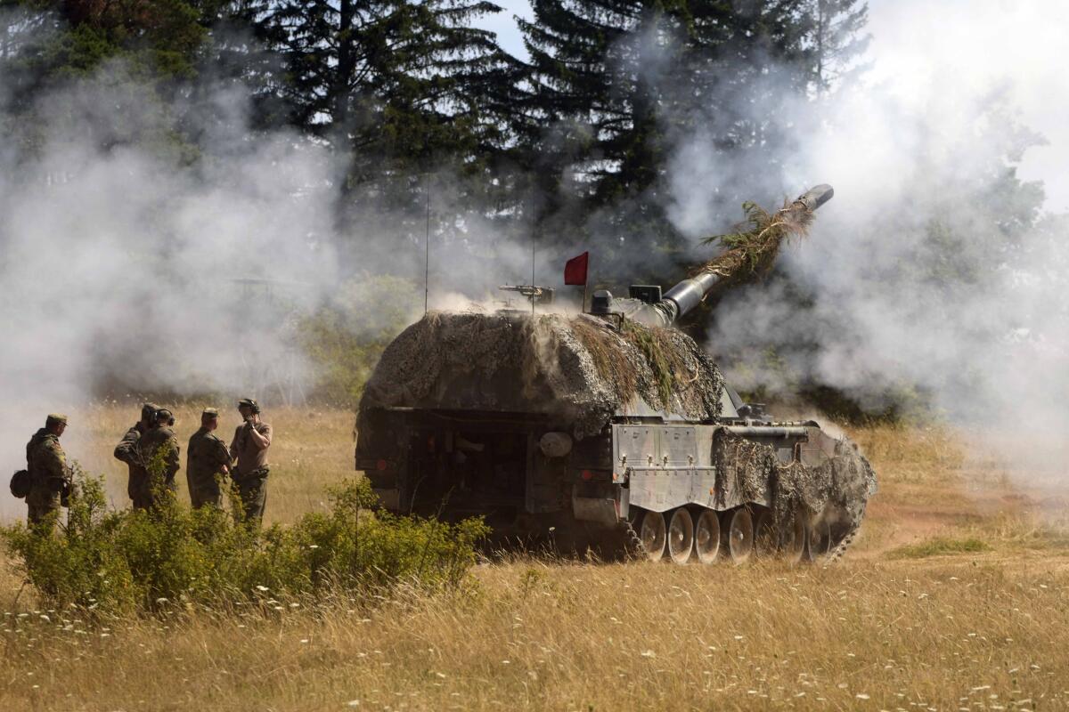 A group of people in camouflage, left, stand near an armored military vehicle as smoke billows near its cannon on a field 