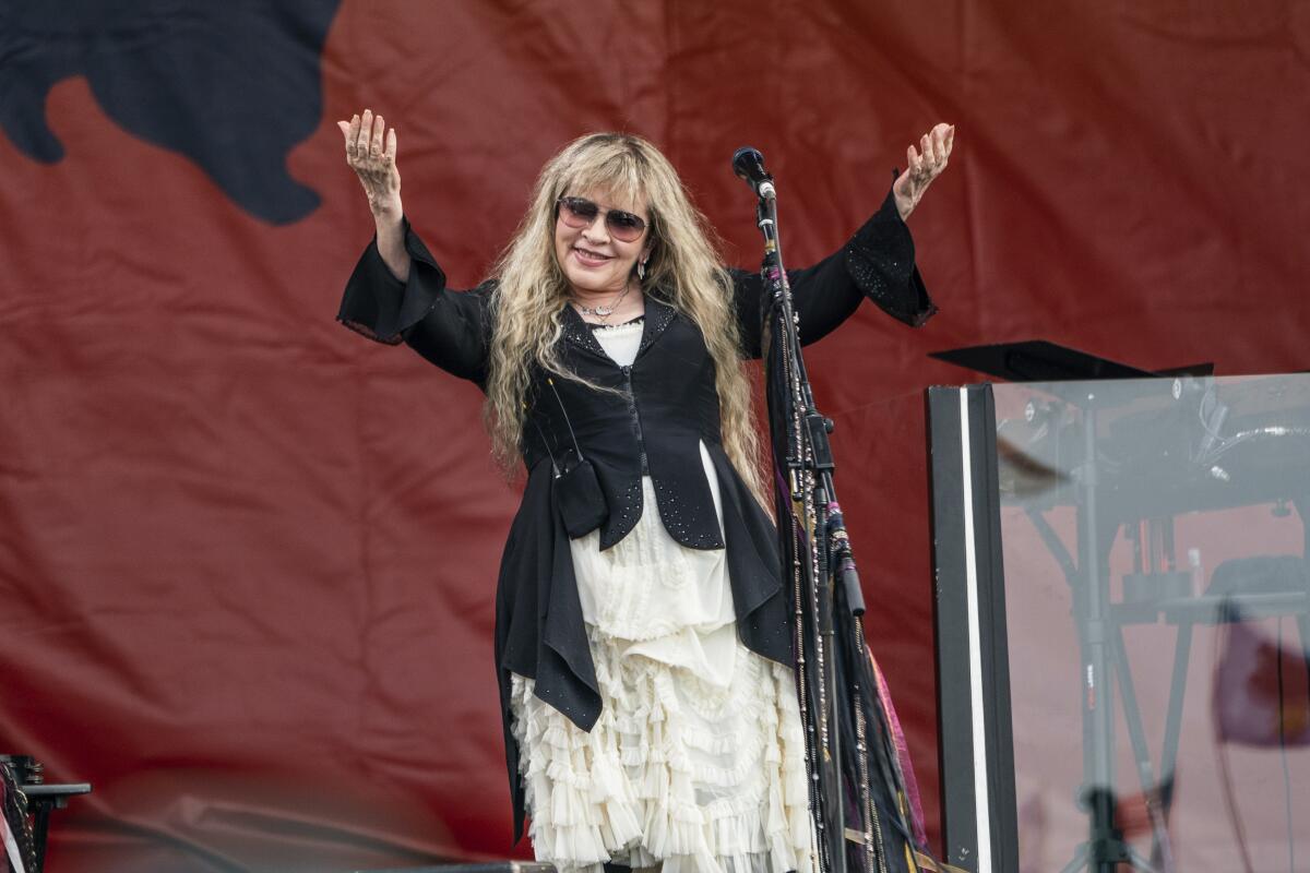 Stevie Nicks on stage in a white tiered skirt and black vest in front of a red backdrop