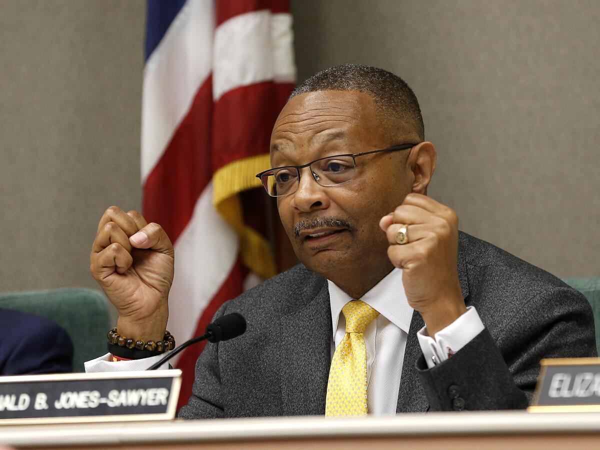 A man in a suit sits on a dais marked with nameplates.