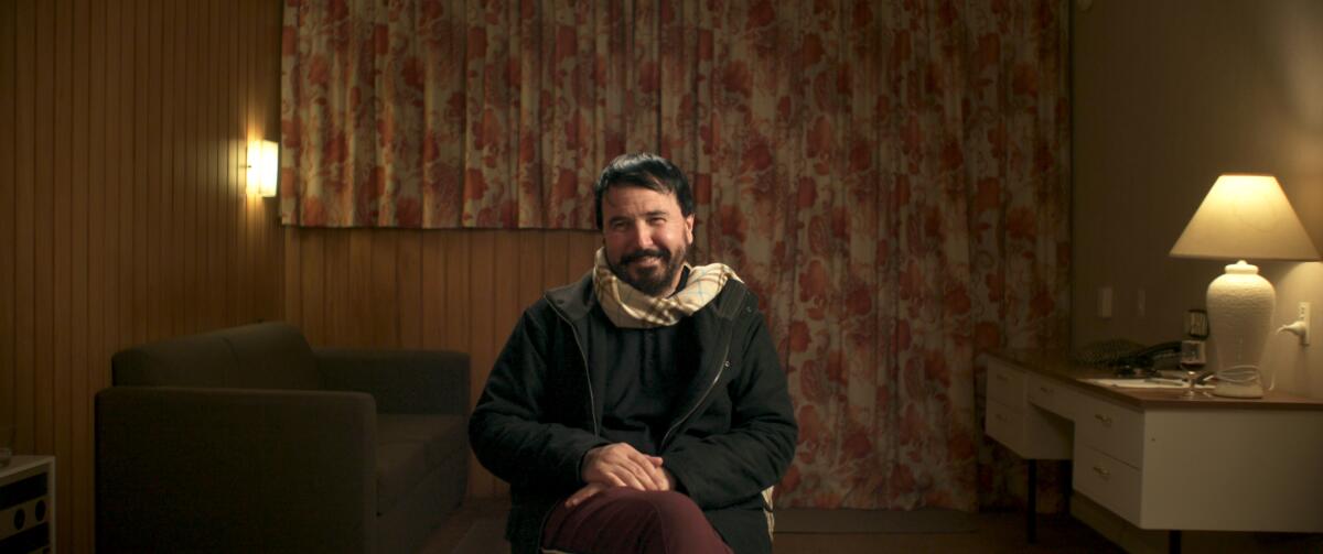 A seated man smiles for the camera in a bland hotel room.