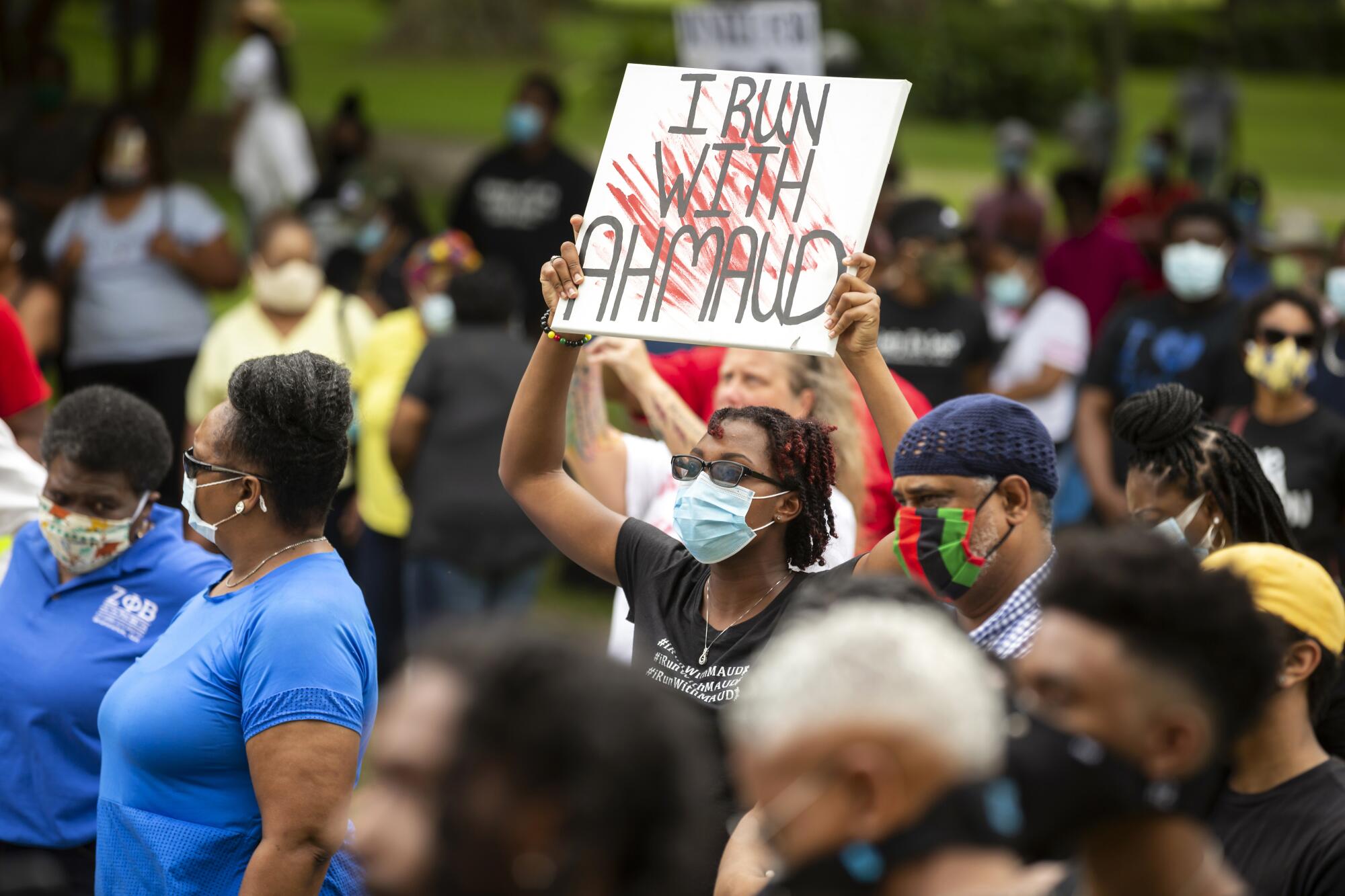 A person holds a sign that says, "I run with Ahmaud," among a crowd of people outdoors.