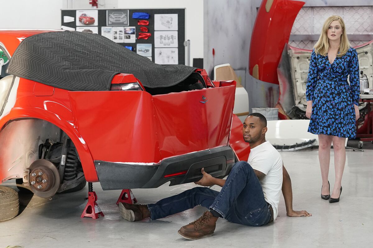 A man sits on the ground next to a car in an auto shop while a woman stands next to him 