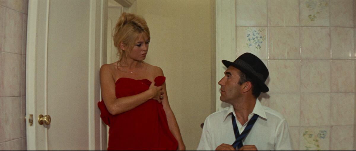 A blond woman wrapped in a red towel looks down at a man in a fedora