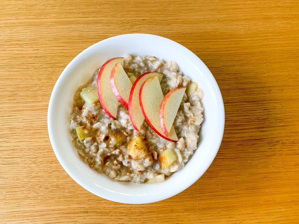 A white china bowl of oatmeal topped with sliced apple rests on a wooden surface.