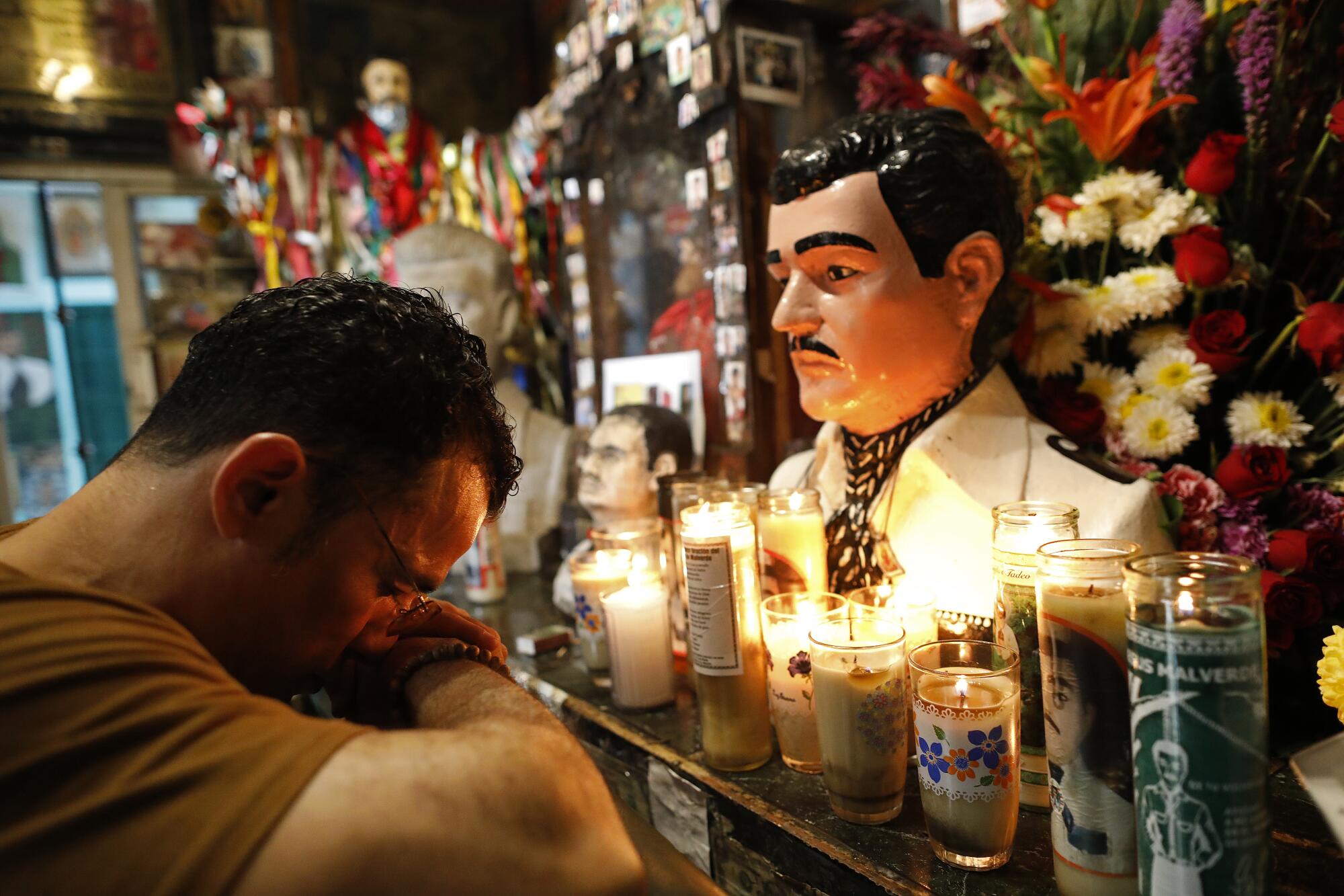 A man bows his head before a bust of Jesus Malverde in a chapel illuminated by candles.