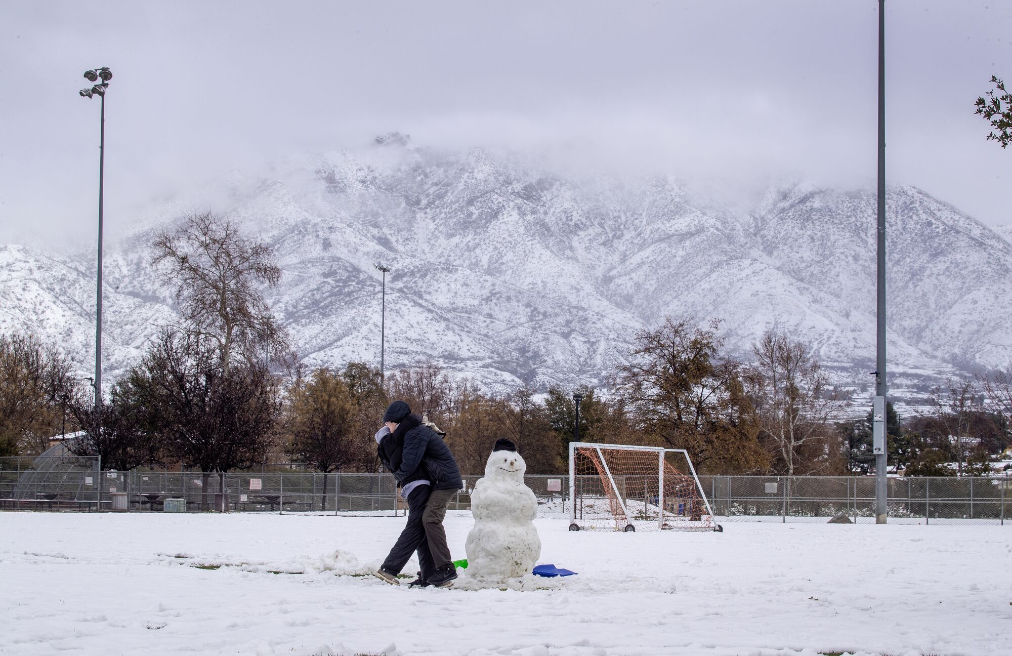 Karla kaplı bir futbol sahasında bir kardan adamın yanında iki kişi kucaklaşıyor