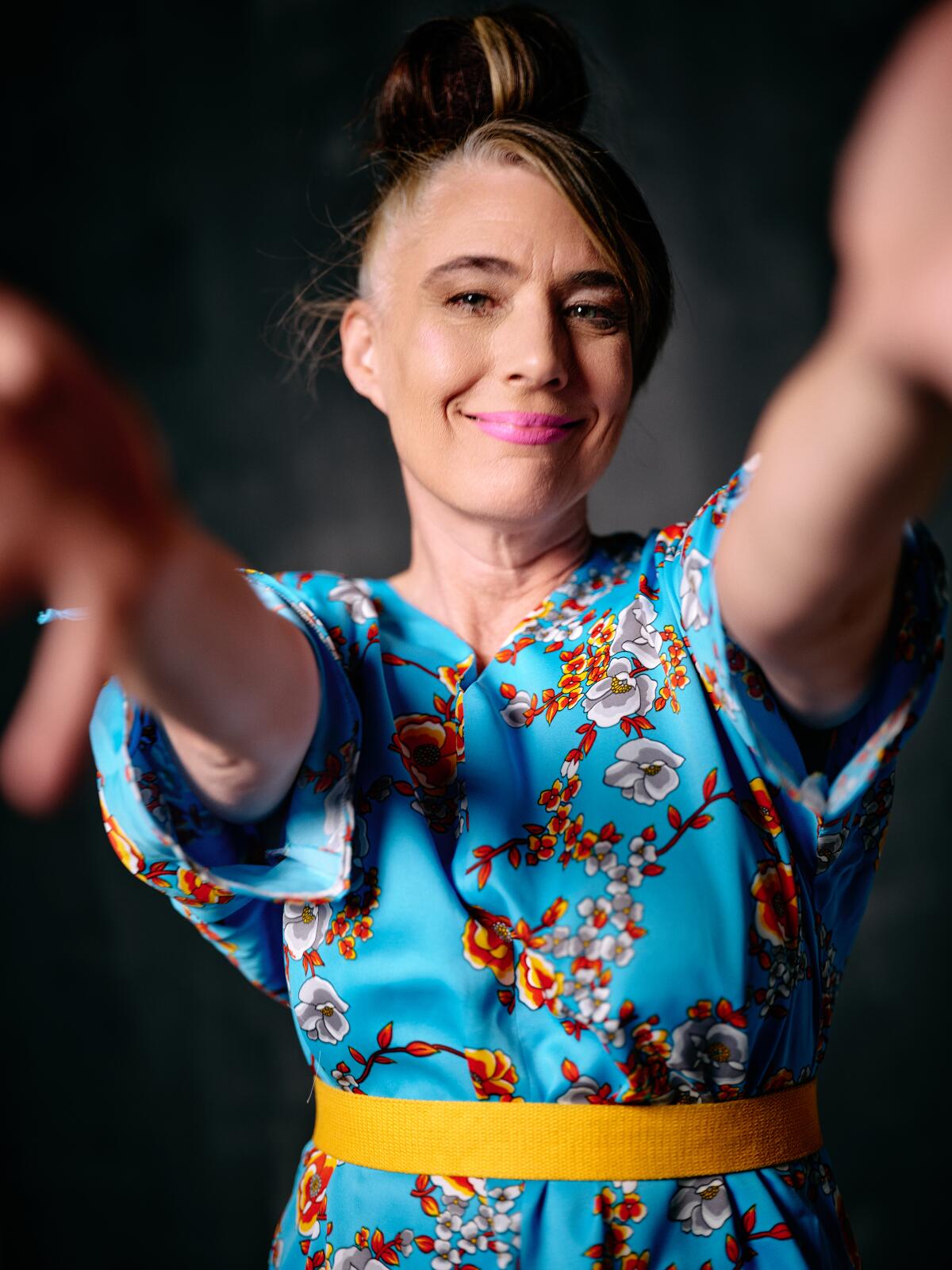 Kathleen Hanna, with pink lipstick, her hair in a bun and wearing a blue floral dress, extends her arms.