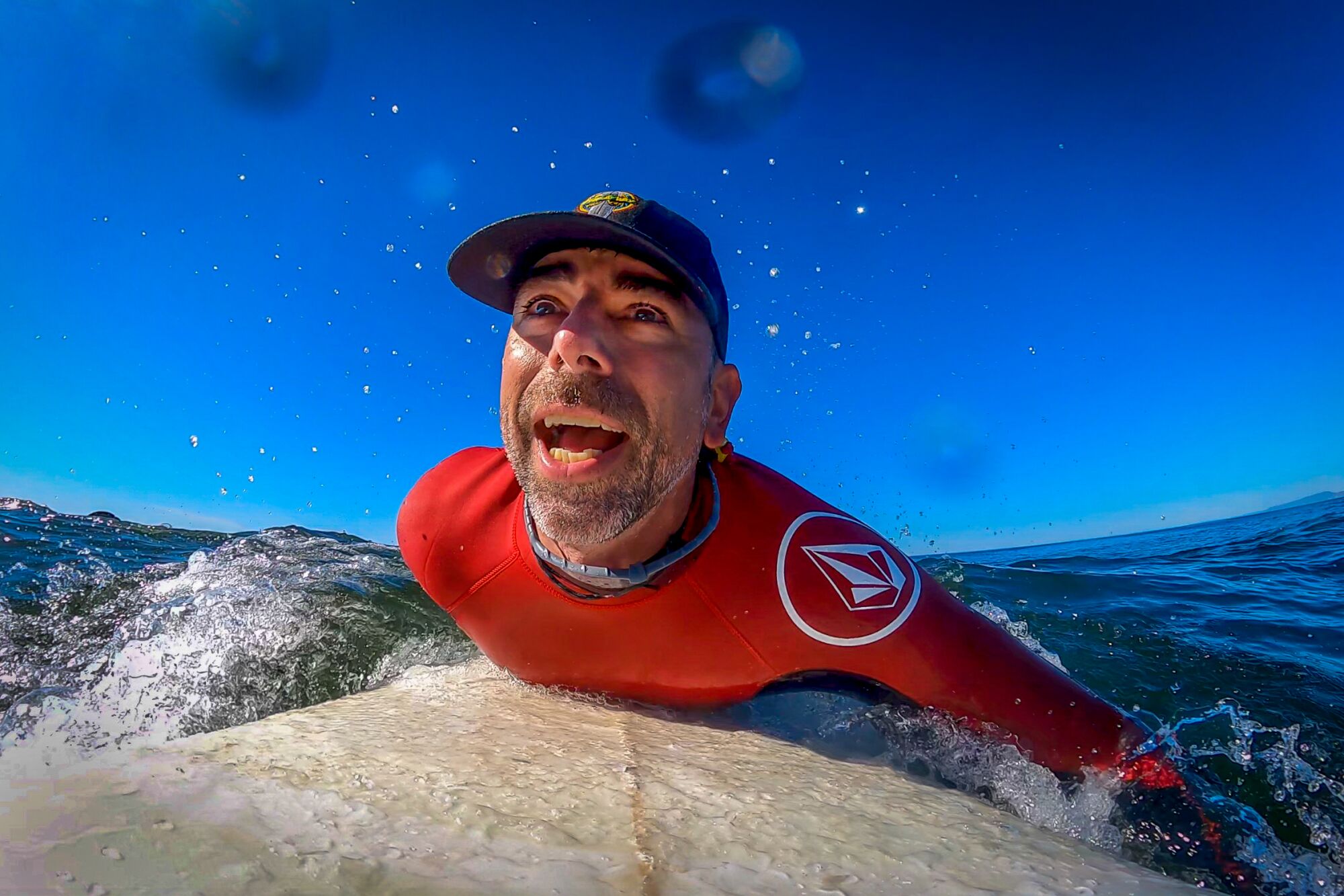 A man in a hat and red wetsuit grins as he paddles his surfboard in the ocean.