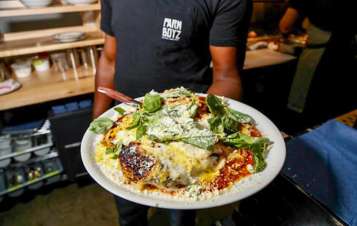 A hand holds out a cheesy, saucy plate of chicken Parmesan from Parm Boyz topped with fresh basil.