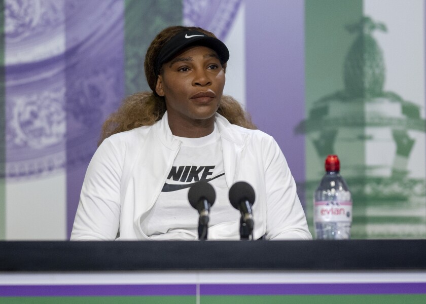 Serena Williams, in a Nike T-shirt and visor, sits behind two microphones with a bottle of water nearby.