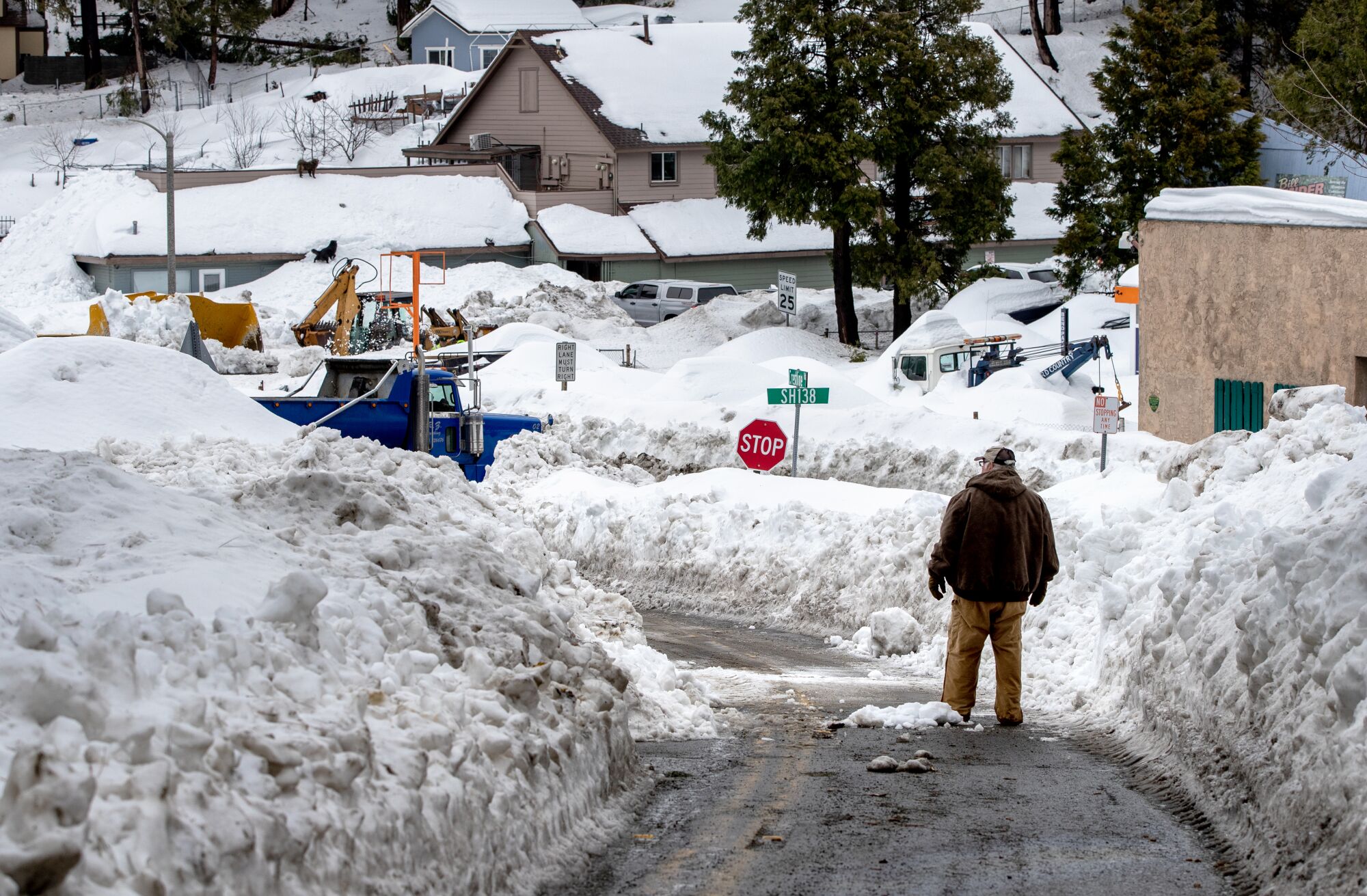 A man stands on a road while piles of snow are stacked up on both sides of it, in some areas higher than the person. 