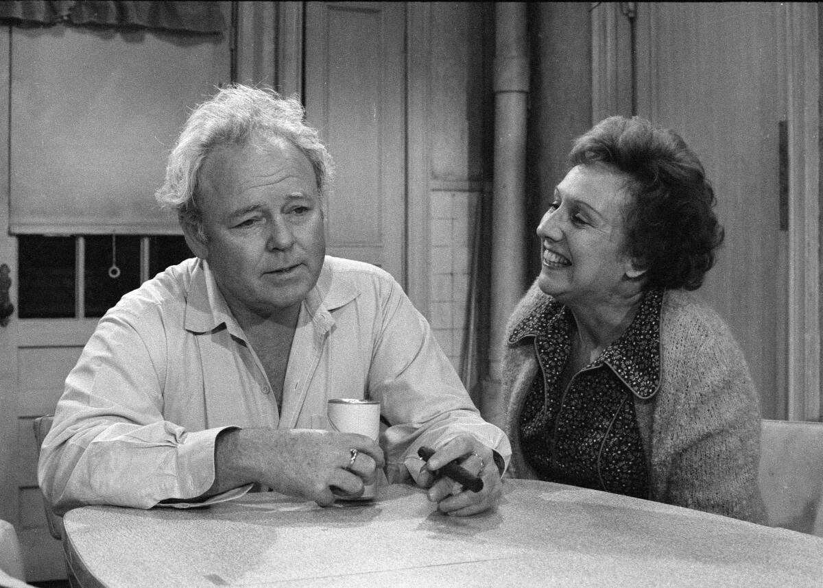A man and a smiling woman sit at a table in "All in the Family." 