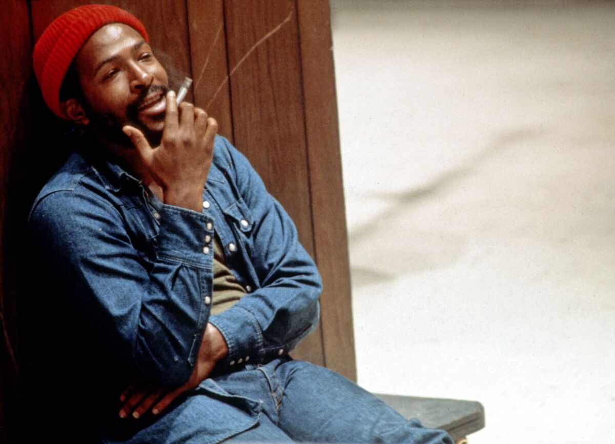 A man in a denim shirt and jeans and a red winter cap smokes a cigarette leaning against a paneled wall.