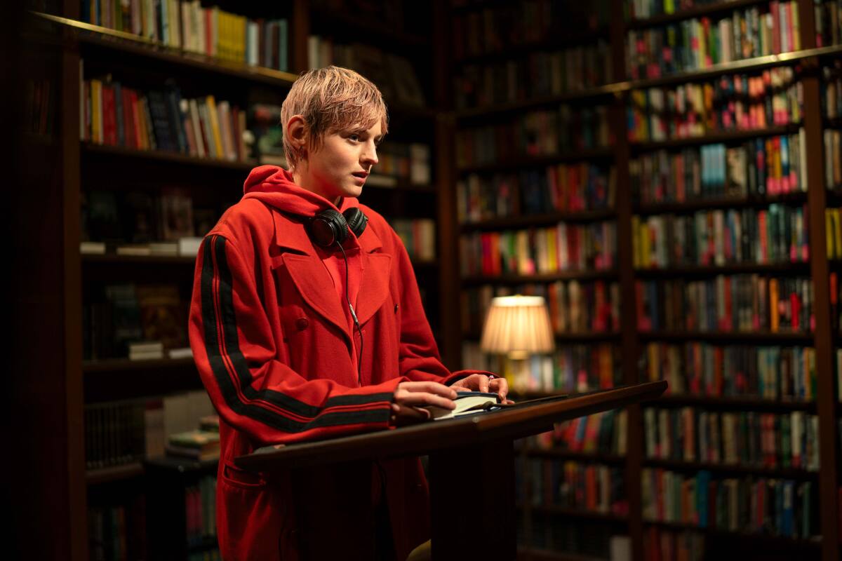 A woman in a red coat with headphones around her neck at a lectern in a bookstore.