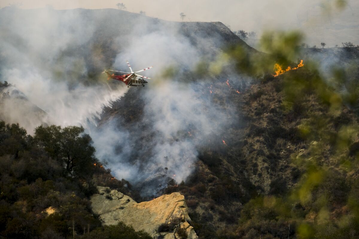 A helicopter flies amid smoke and scattered flames on a rocky hillside.