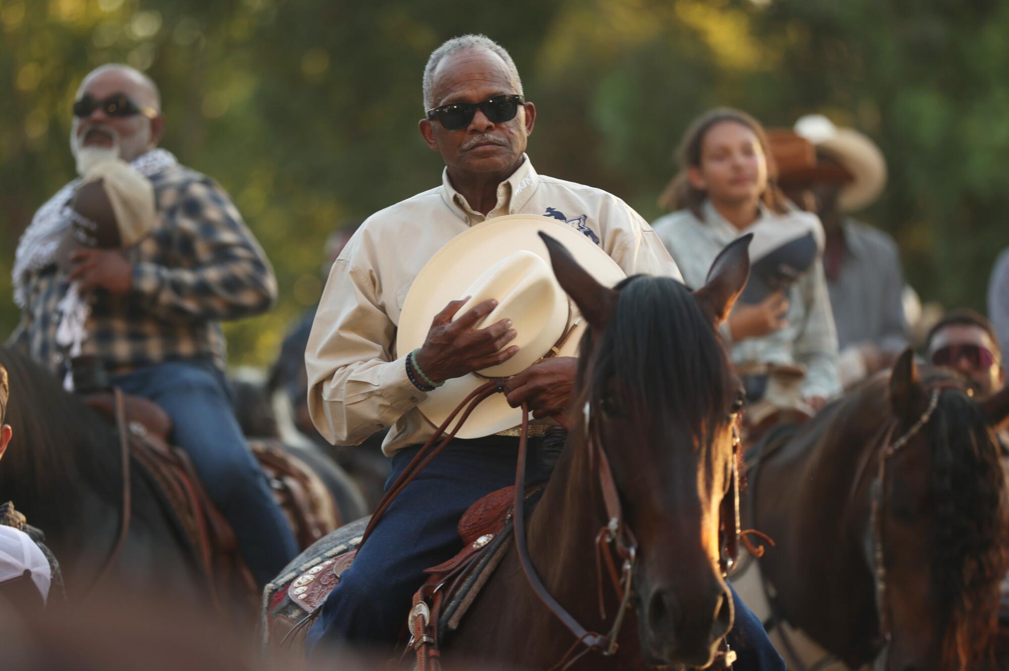 Riders on horses hold their hats against their chests.