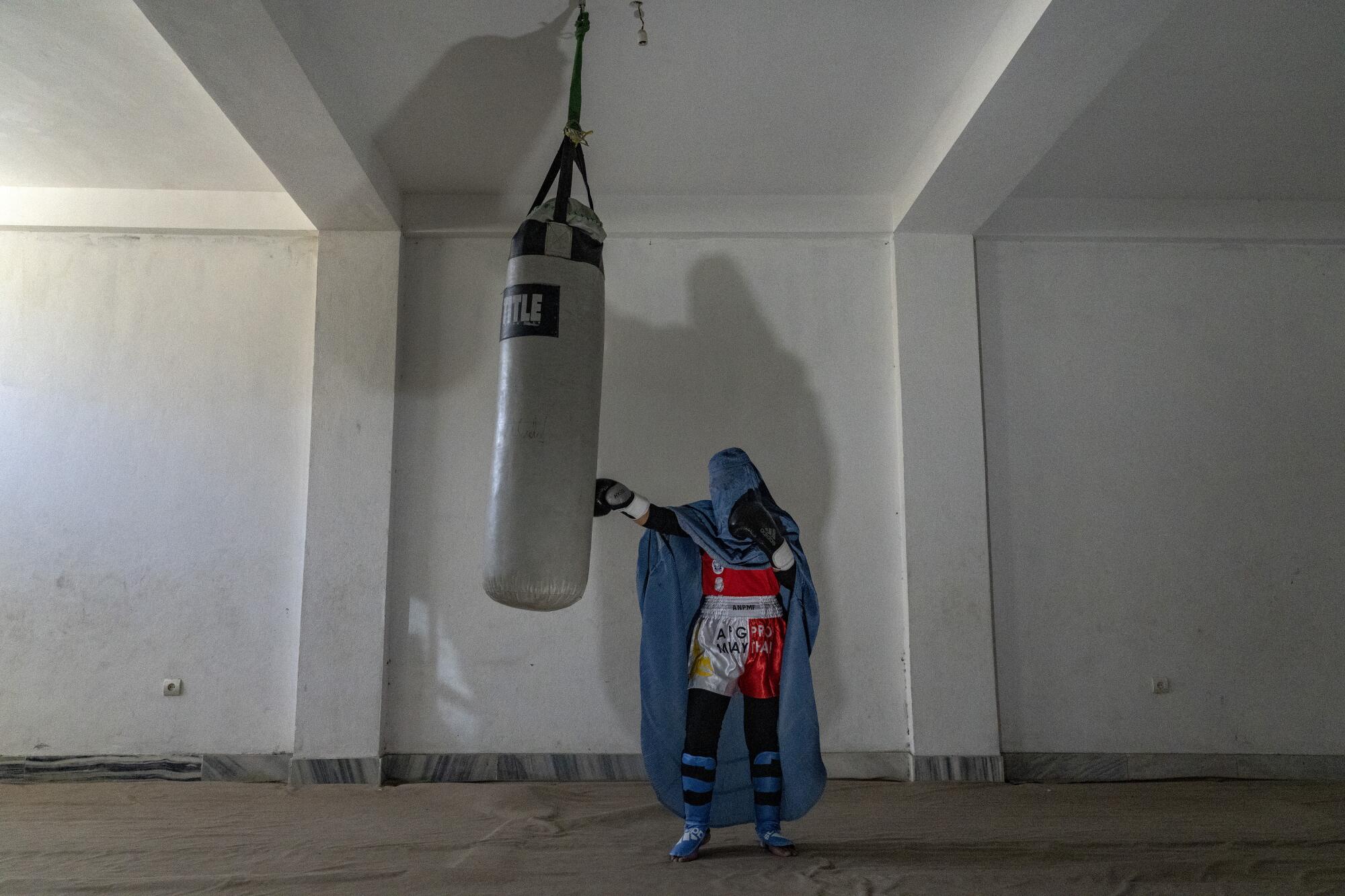 A woman wearing a burqa and boxing gloves punches a workout bag.