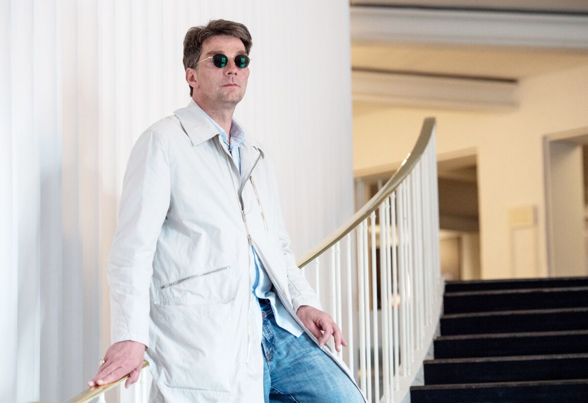 A man in a long jacket and small round sunglasses leans on the railing of a curving inside staircase