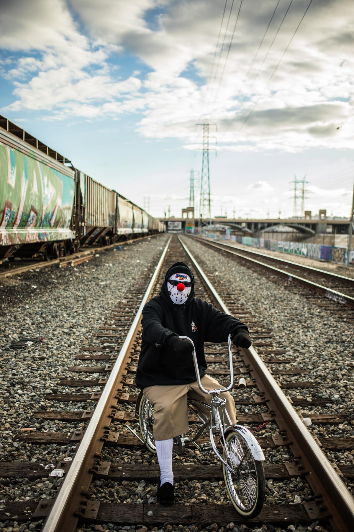 A man wearing a hoodie and mask on a bike on train tracks