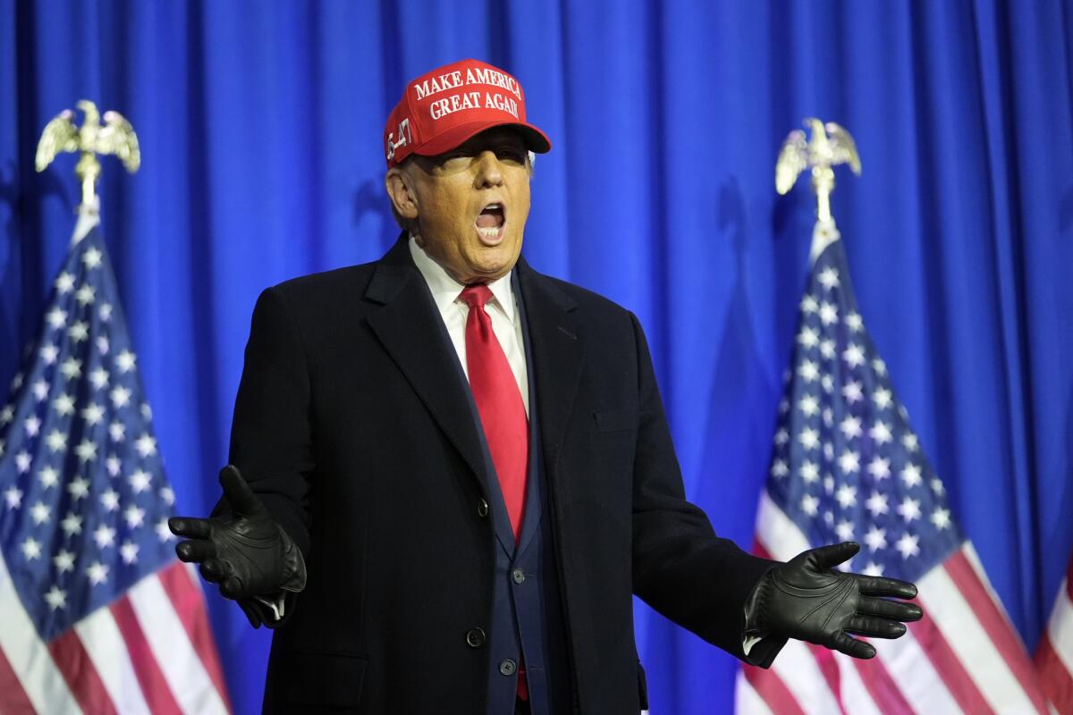 Ex-President Trump, in black gloves and a MAGA hat, speaking in front of a royal-blue stage curtain and two American flags