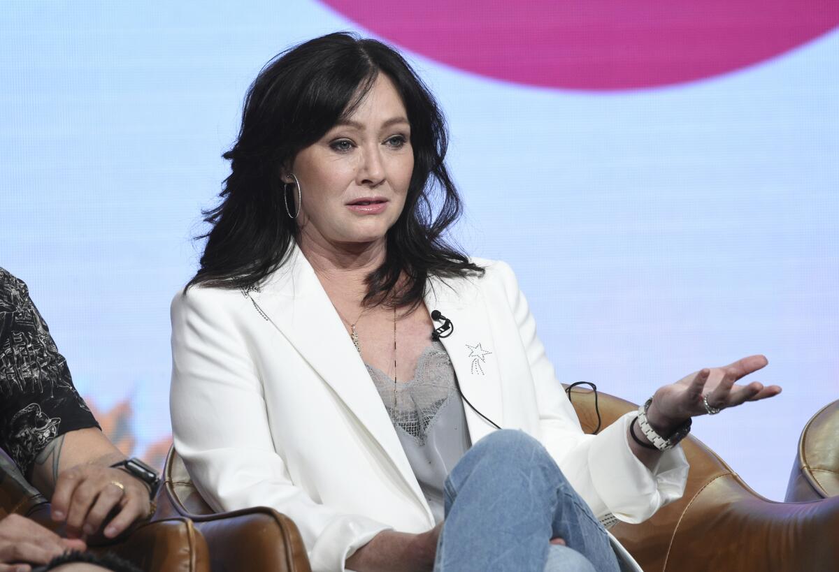 Shannen Doherty wears a white blazer over a gray blouse with jeans and sits in a chair speaking. 