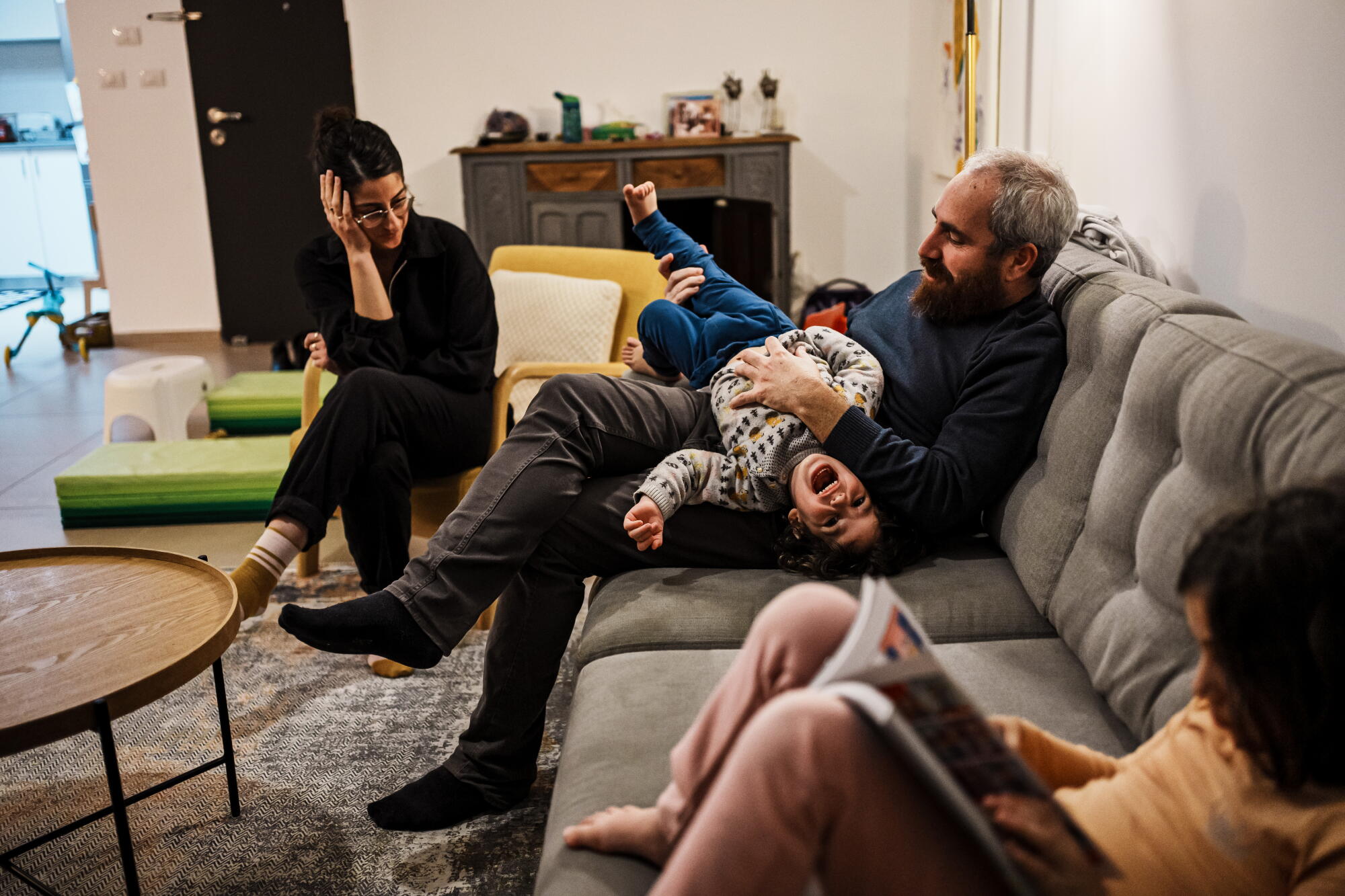 A man seated on a couch holds a child in his lap, flanked by a woman and an older child, who is reading a book
